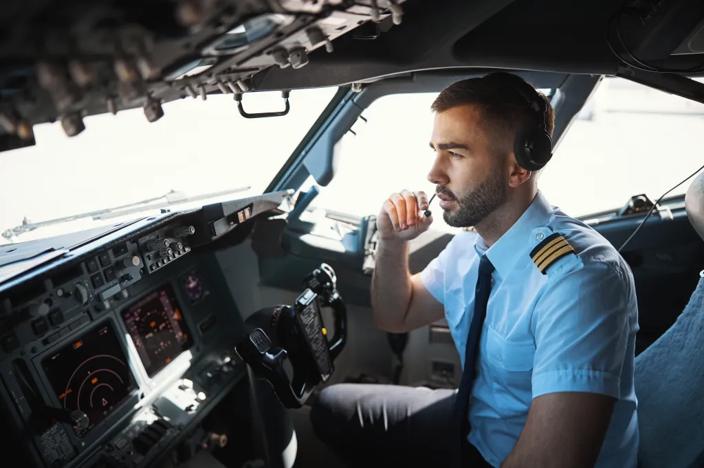 A young pilot in a white shirt with a tie and epaulets sits in an airplane cockpit, communicating via a transmitter microphone, with the dashboard and pilot's controls visible in the foreground.