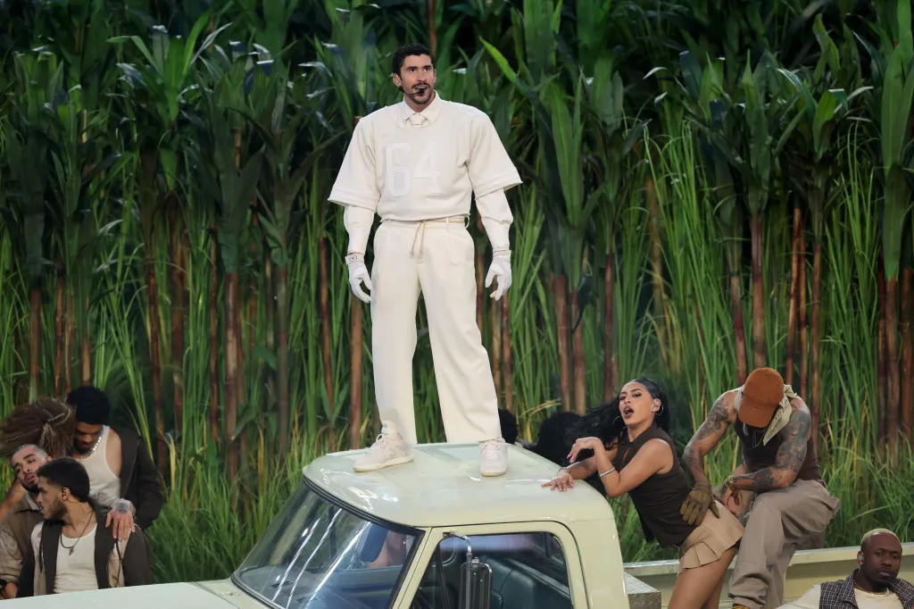 Bad Bunny performs during the Super Bowl halftime show, standing on a light green vehicle with backup dancers around him, in front of tall green plants.