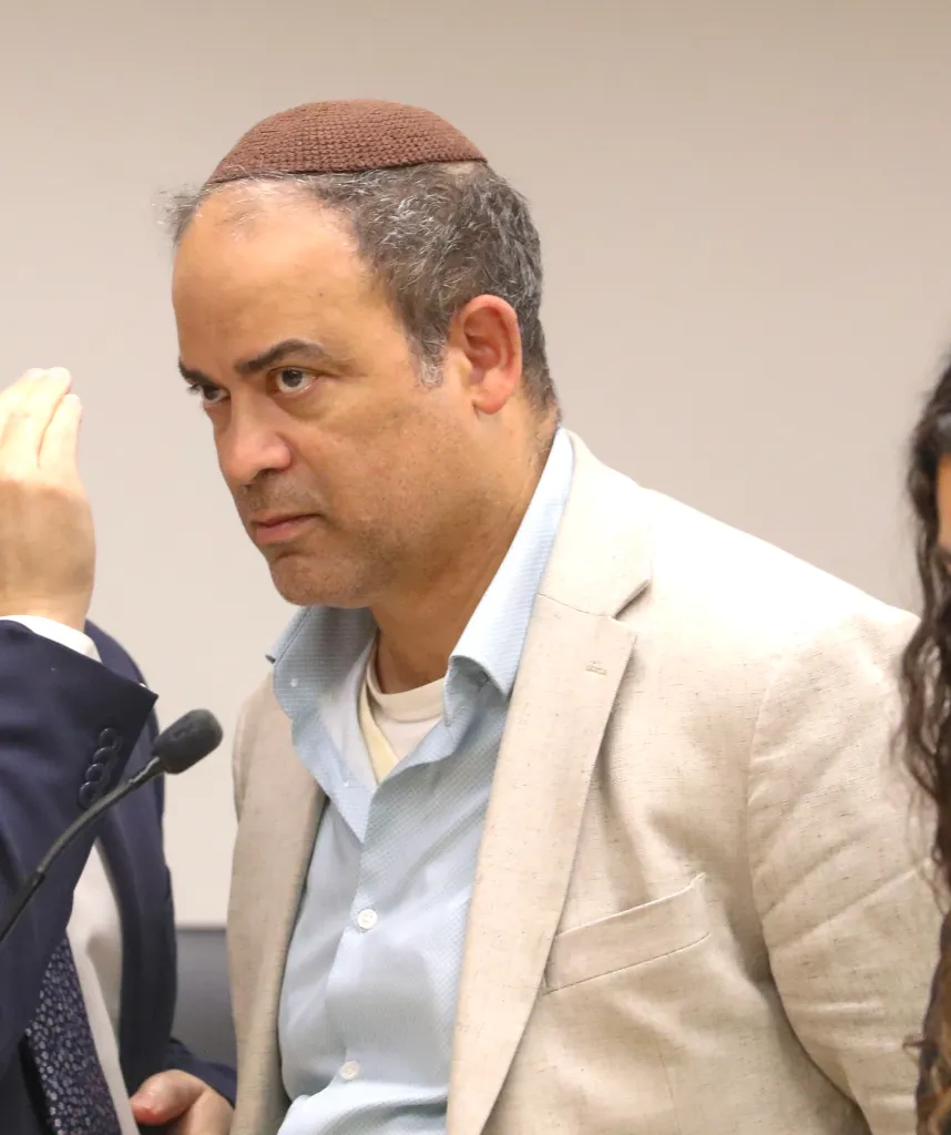 A man with a brown yarmulke, light blue shirt, and beige jacket is shown from the chest up, looking to his left.