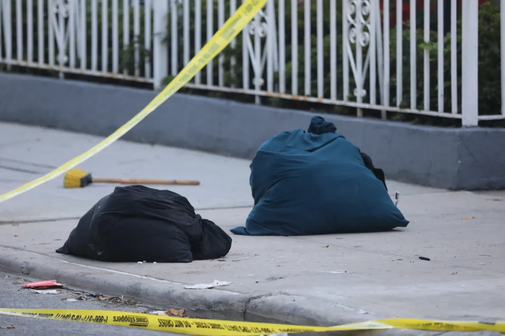 Two body bags lie on the sidewalk at a stabbing scene, with yellow crime scene tape visible.