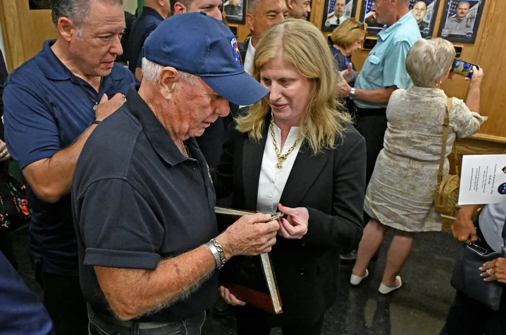 A man in a blue baseball cap hands a woman in a black blazer and white shirt a small, engraved item.