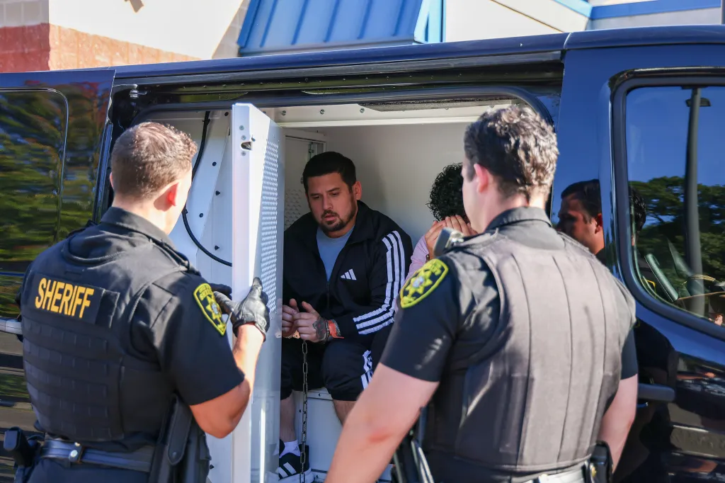 Sage Arias-Brown, in handcuffs, sits in the back of a police van as two sheriff's deputies stand nearby.