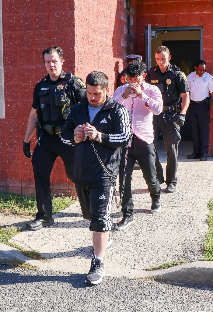 Sage Arias-Brown in Adidas track suit, chained to another man in a pink shirt, being led by two officers with a third officer behind them.