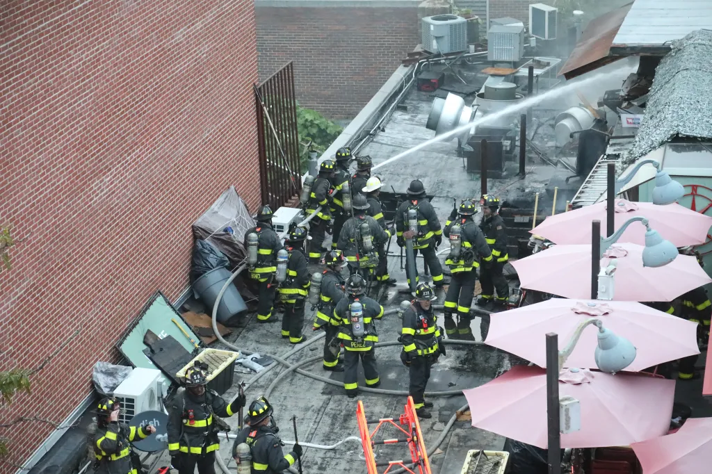 Firefighters on a rooftop spray water on a fire, with pink patio umbrellas visible.