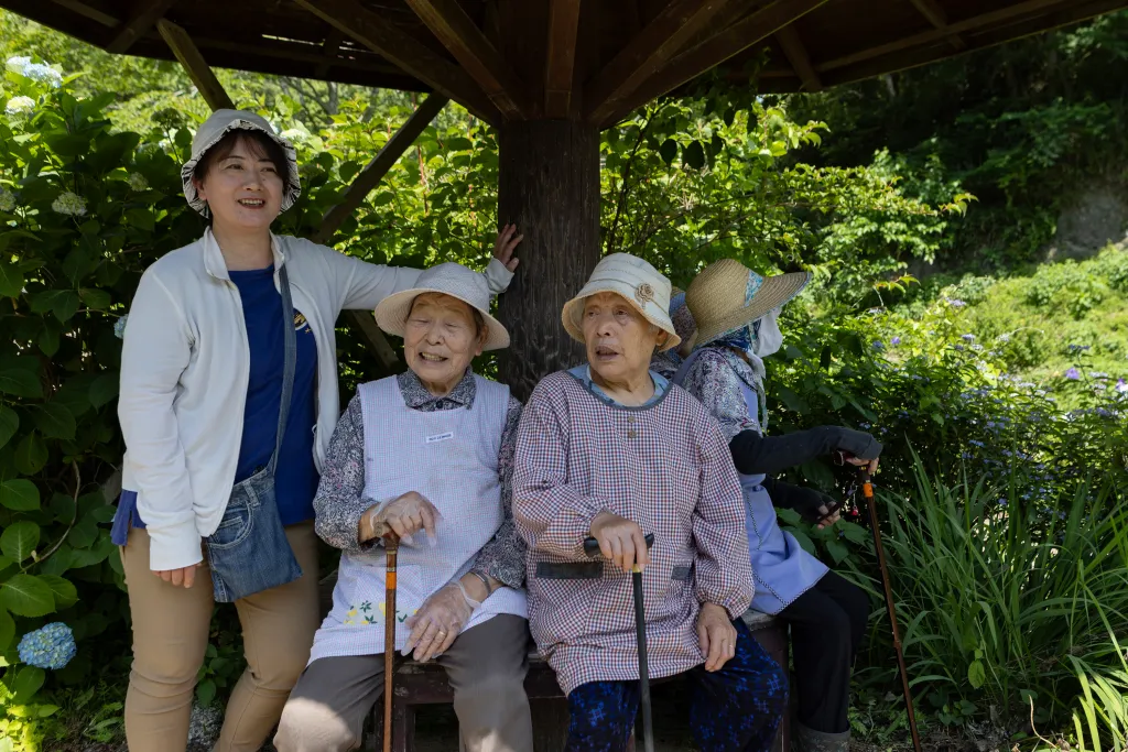 A group of three elderly women and one younger woman resting under a wooden structure amidst lush greenery.
