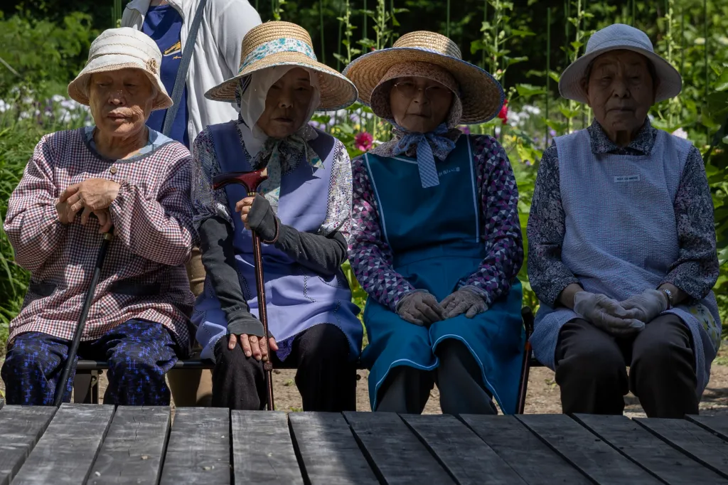Four elderly Japanese residents, wearing hats and aprons, sit on a bench in front of a flower garden.