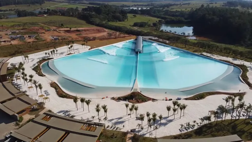 An aerial view of the Wavegarden Cove surf lagoon, featuring a large, man-made pool with white sandy beaches and palm trees, surrounded by green landscape.