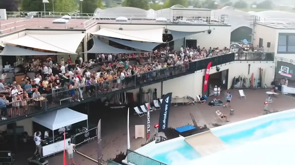 Aerial view of a surf park with a large crowd gathered on an outdoor balcony overlooking a wave pool.