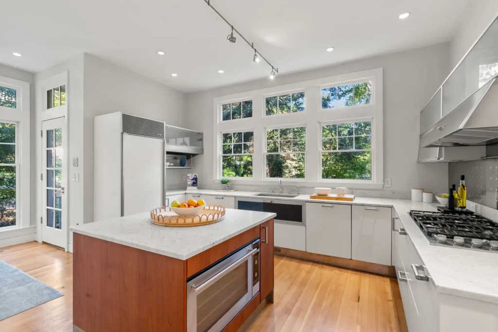 Kitchen with a white center island, white cabinets, stainless steel appliances, and large windows looking out to trees.