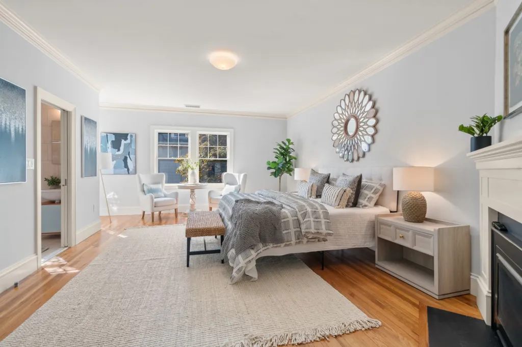 The primary bedroom in Julia Child's former home has light blue walls, hardwood floors, a large rug, a queen bed, and a sitting area by the window.