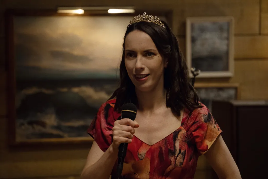 Kate O'Flynn as Kate in Widow's Bay, wearing a tiara and holding a microphone.