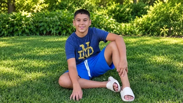 A boy sits on green grass with his legs bent, wearing a blue shirt with a yellow logo, blue shorts, and white slides.