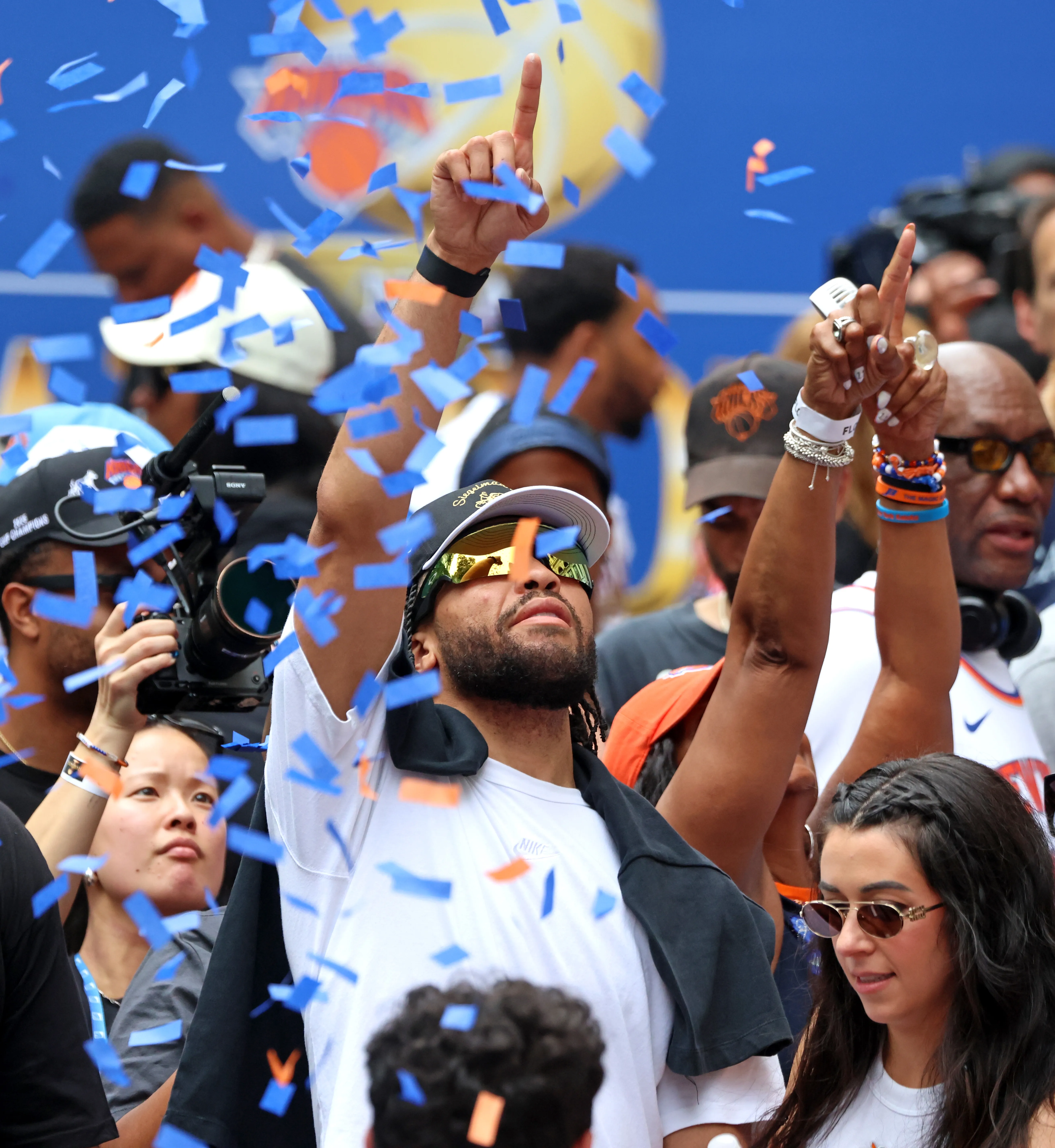 New York Knicks player Jalen Brunson celebrating on a parade float as blue and orange confetti falls around him.