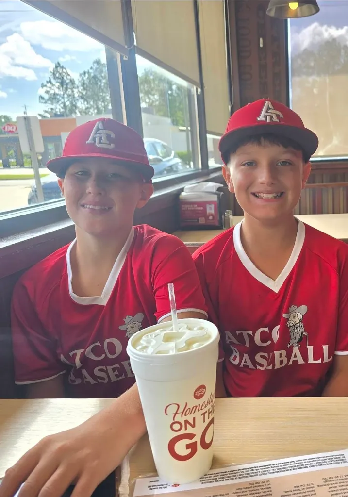 Two smiling boys in red baseball jerseys and hats sit at a table in a restaurant.
