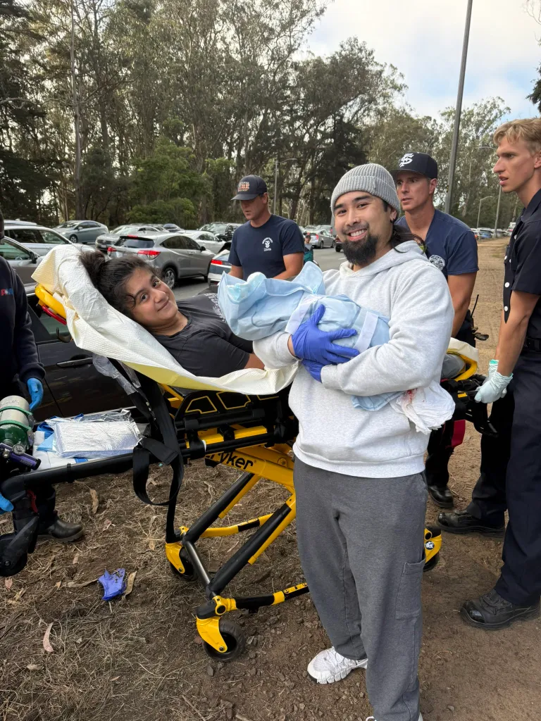 A man holds a newborn baby while standing next to a woman on a stretcher, with emergency responders in the background.