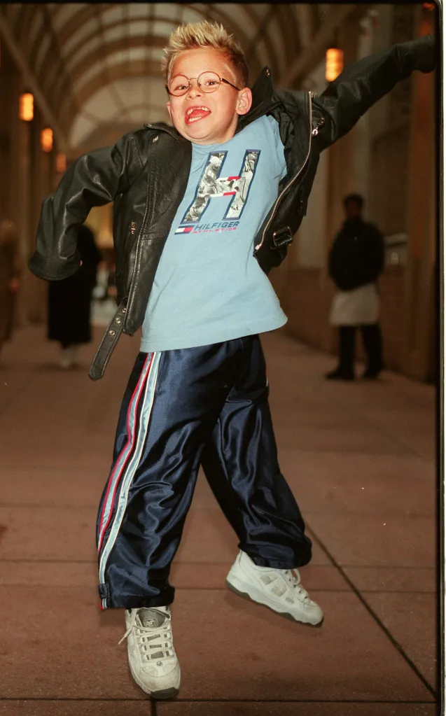 Actor Jonathan Lipnicki wearing a leather jacket and glasses jumps enthusiastically in a hallway.