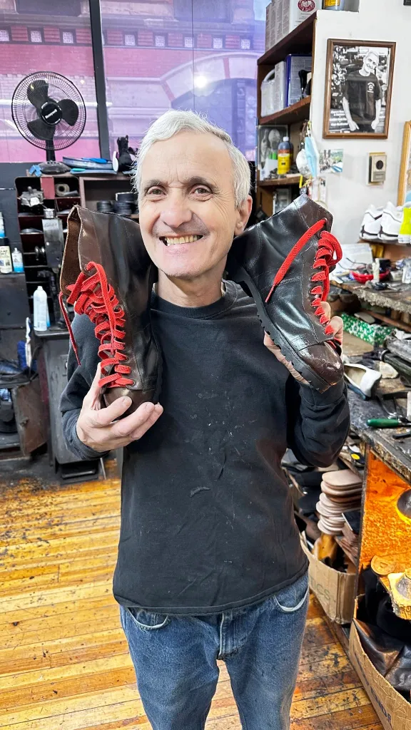 A man in a shoe repair shop holds up a pair of boots with red laces.