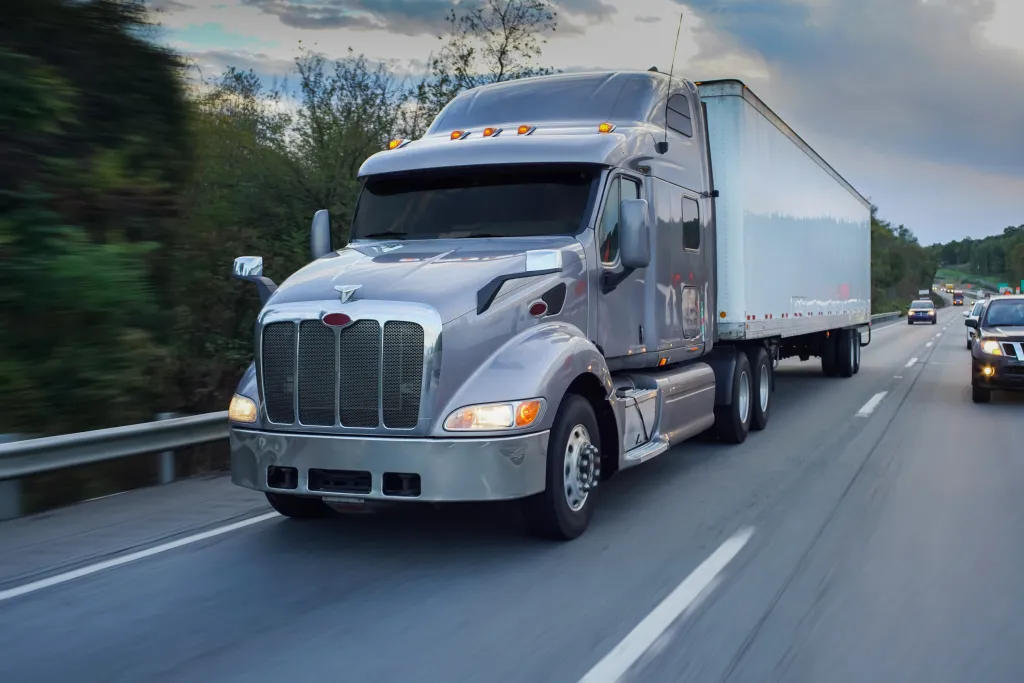 A gray 18-wheeler semi-truck with a white trailer drives on a highway next to blurred trees.