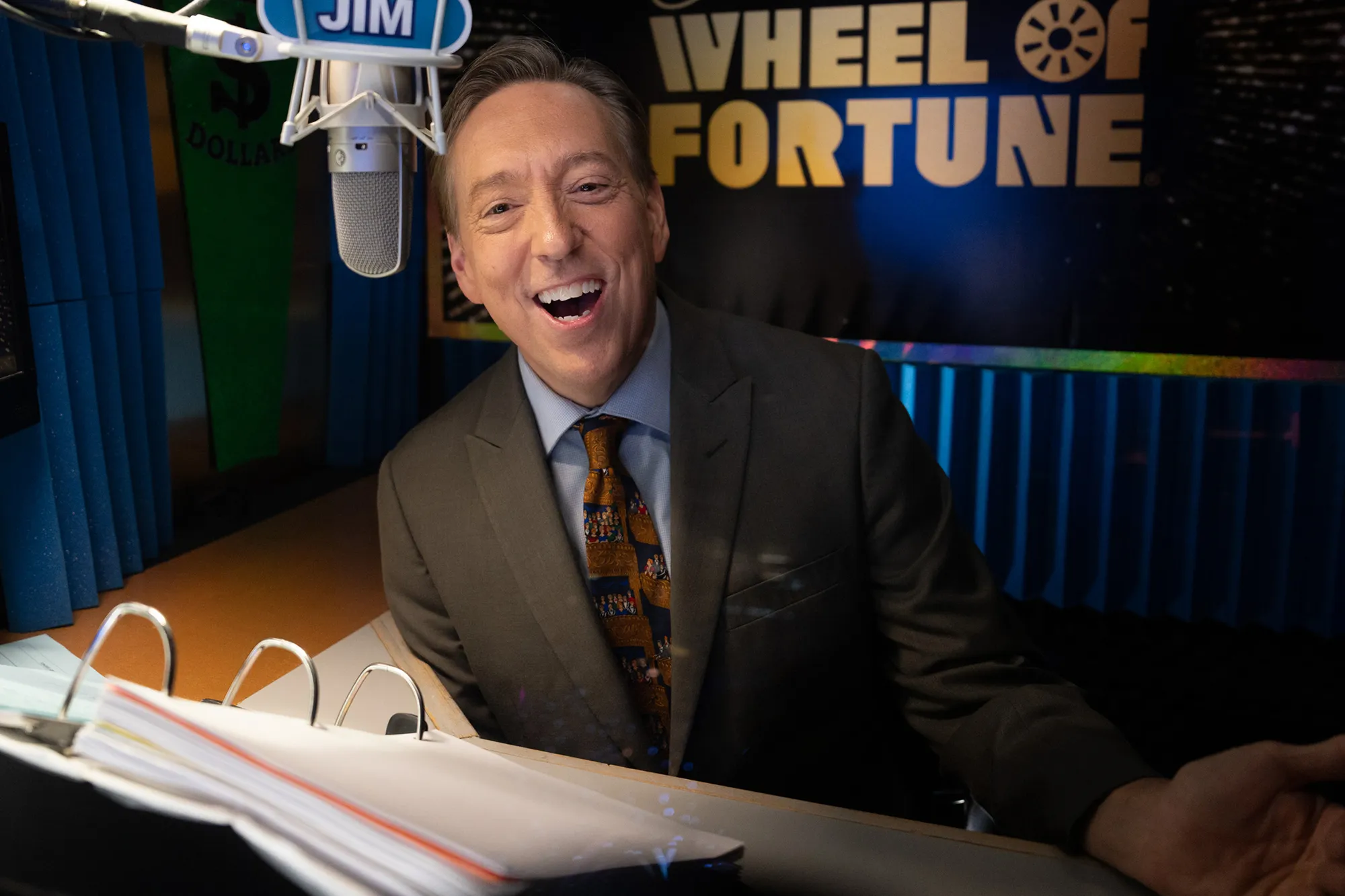 A man with an open-mouthed smile, wearing a suit and tie, sitting in a recording booth labeled 