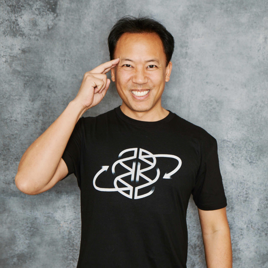 A man in a black t-shirt with a white circular logo pointing to his temple and smiling.