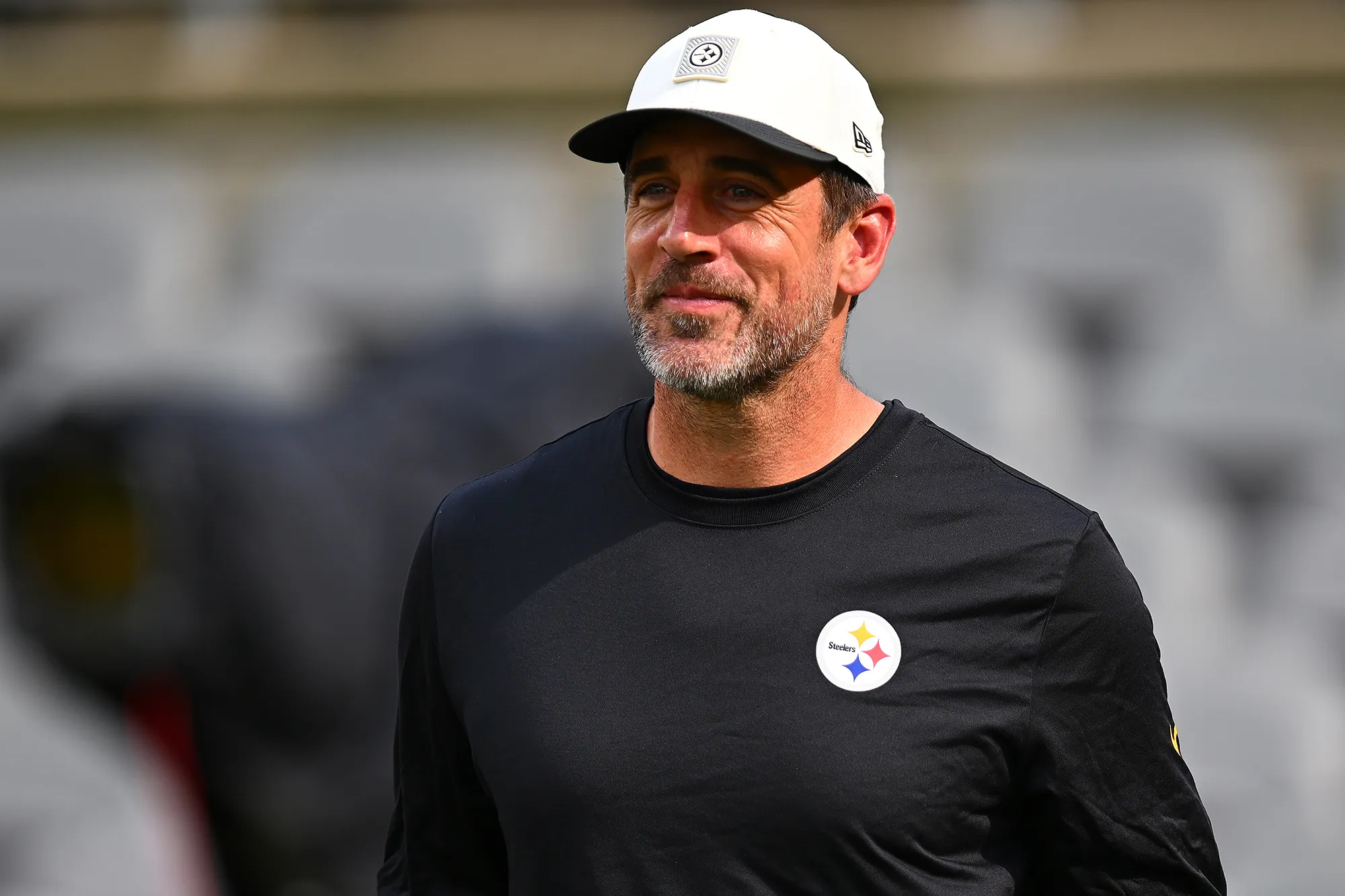 Aaron Rodgers in a black shirt with a Pittsburgh Steelers logo and a white baseball cap.
