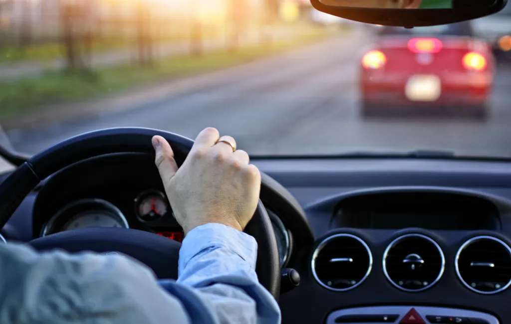 Person driving a car from the driver's perspective, with a red car visible through the windshield.