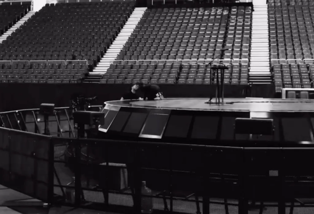 A person crawling on a circular stage in an empty arena.