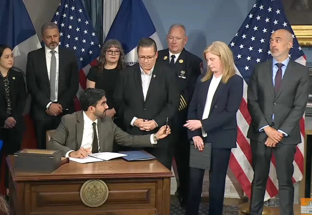 A man signing a document at a desk, surrounded by other officials.