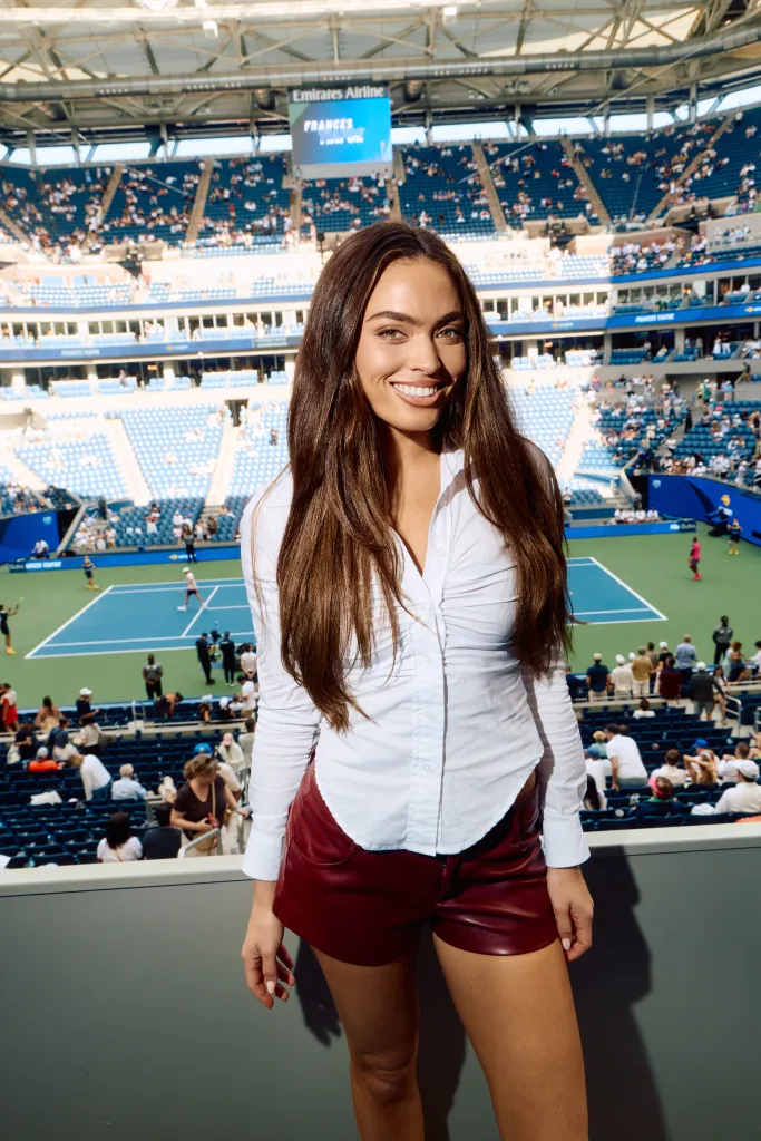 Grace Ann Nader in a white shirt and red shorts smiles at the camera from a box seat overlooking a tennis court.