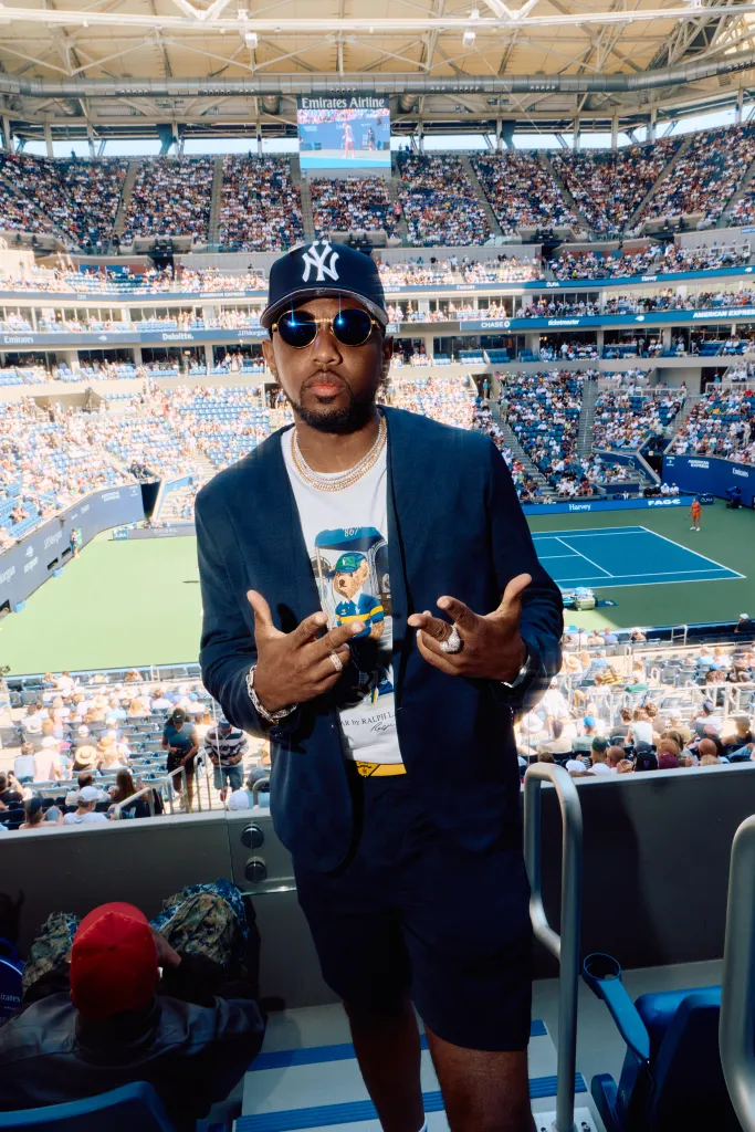 Fabolous in a Yankees cap and sunglasses poses in a stadium with a tennis court and spectators visible in the background.
