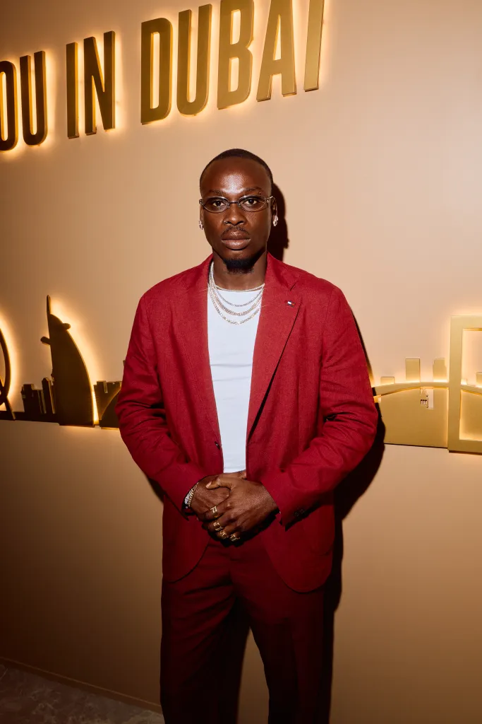 Fireboy DML in a red suit, white shirt, and multiple diamond chains and rings posing in front of an illuminated 