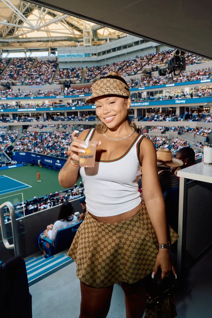 Latto wearing a white tank top, a patterned Gucci visor, and a matching skirt poses at the US Open, holding a drink.