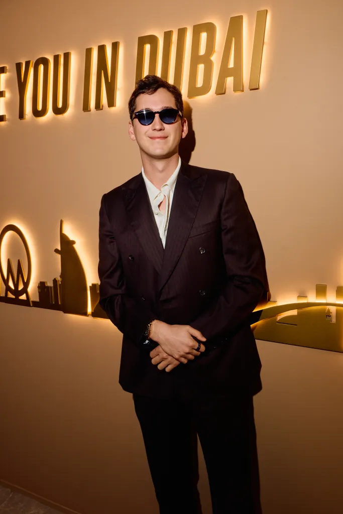 A man wearing sunglasses, a double-breasted suit, and a light-colored collared shirt stands smiling in front of a lighted sign that says 