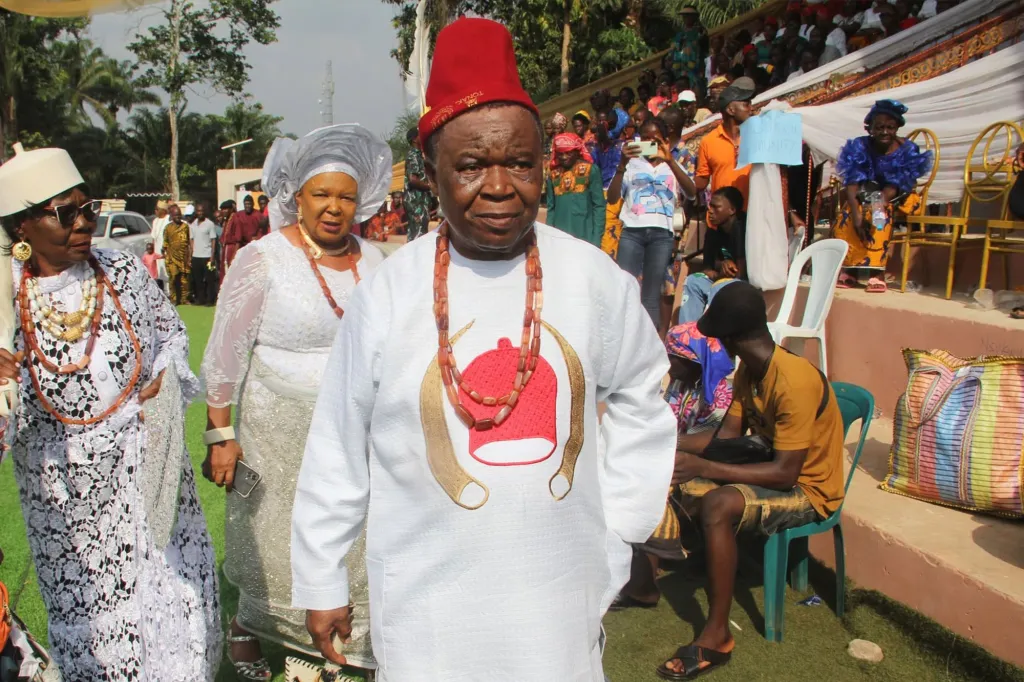 A man in a red hat and white tunic adorned with beads, flanked by two women in white dresses, stands amidst a gathering.