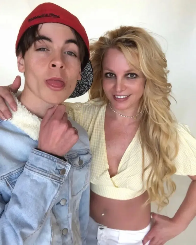 A man in a red baseball cap and denim jacket, sticking out his tongue, stands next to Britney Spears.