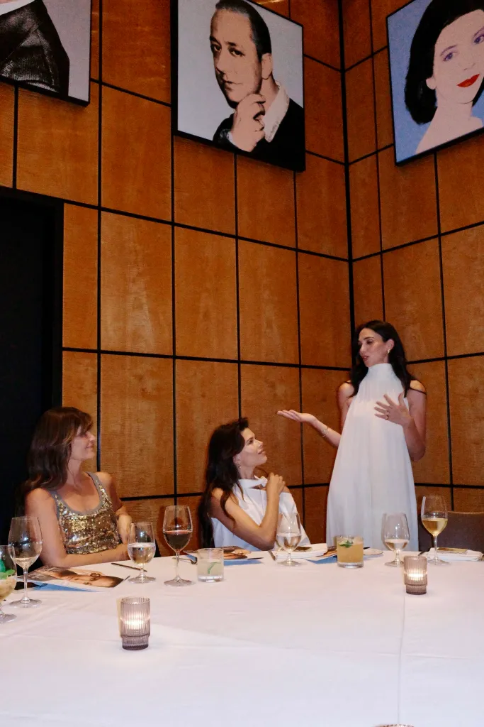 A woman in a white dress stands addressing two women seated at a table, with portraits on the wall behind them.