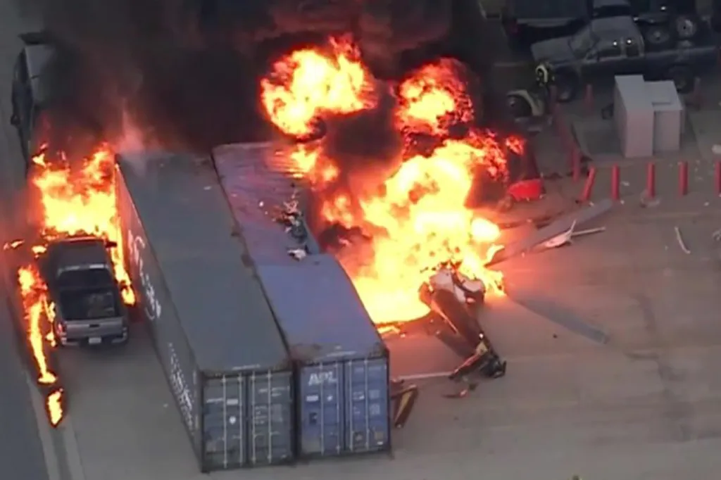 Aerial view of multiple vehicles and shipping containers engulfed in flames with thick black smoke.