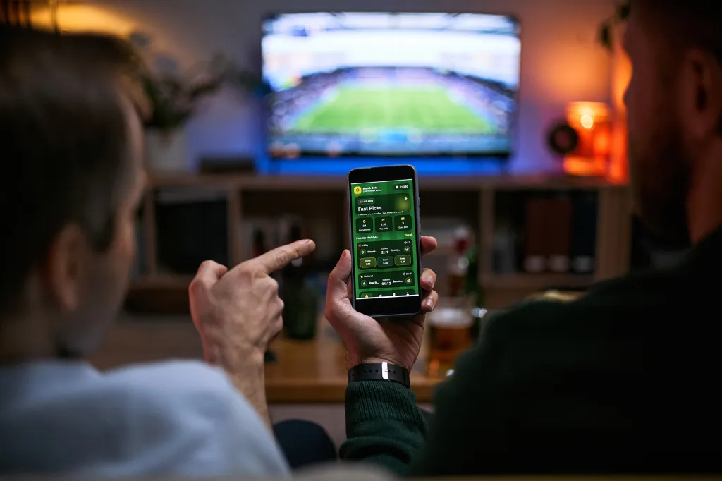 Two men watching a football match on TV and using a sports betting app on a smartphone.