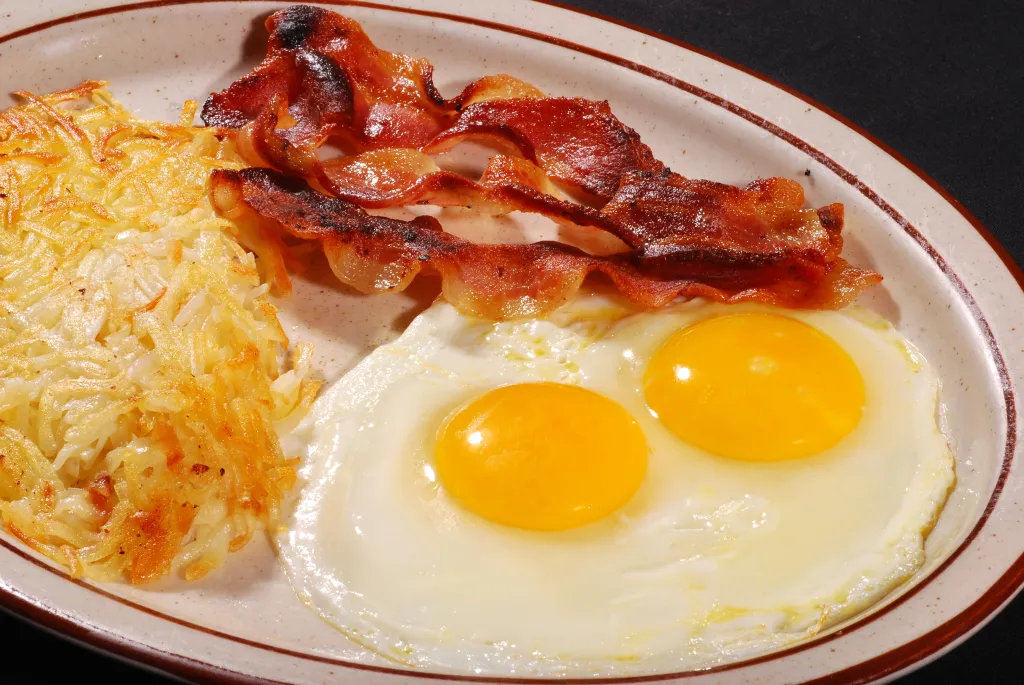Breakfast plate with hash browns, crispy bacon strips, and two fried eggs with bright yellow yolks.