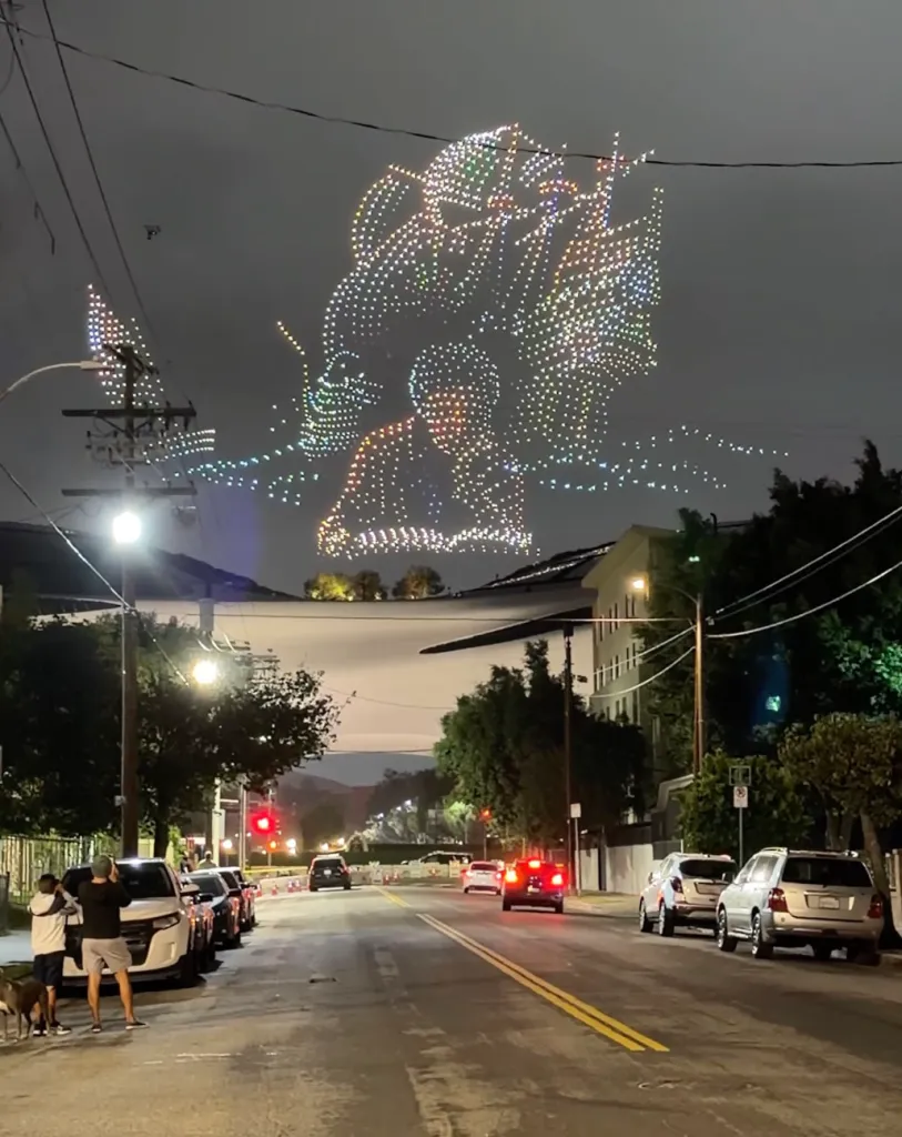 Drone light show depicting a figure and an abstract shape in the night sky over a street with cars and onlookers.