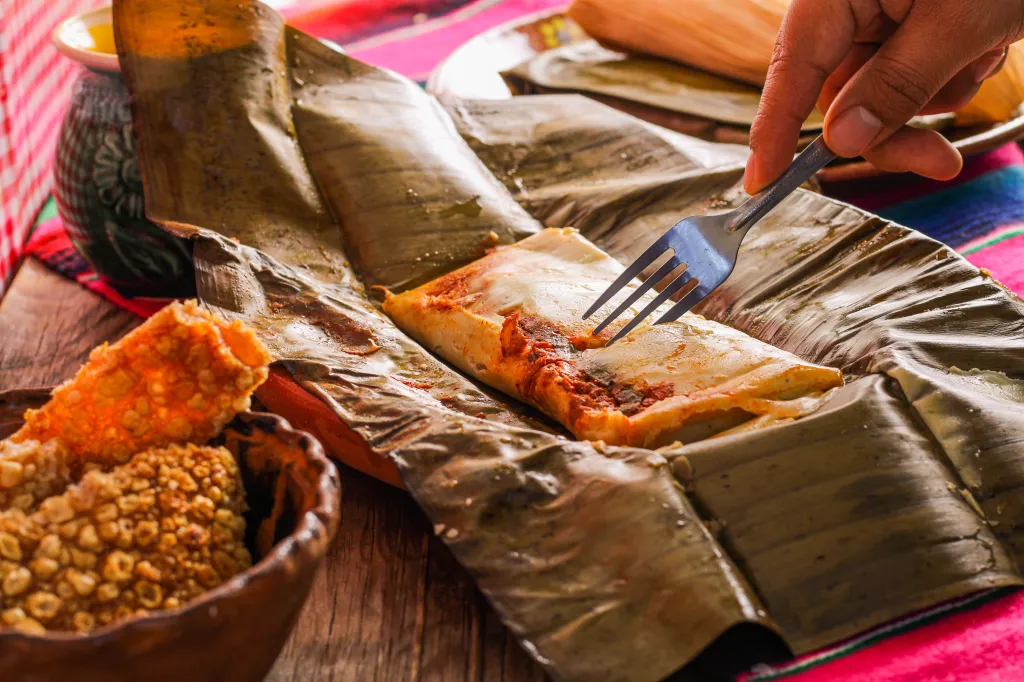 A hand uses a fork to open a Veracruz tamal, revealing its filling of pork and red sauce, next to a bowl of chicharrón.