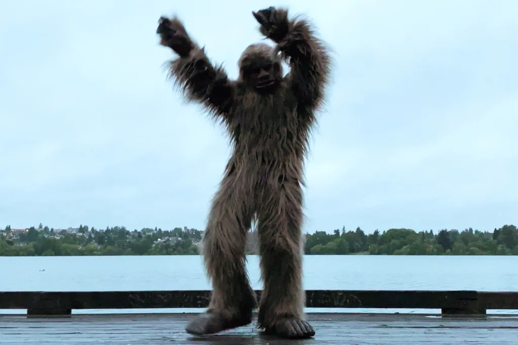 A person in a Bigfoot costume on a pier with a lake and distant shoreline behind them.