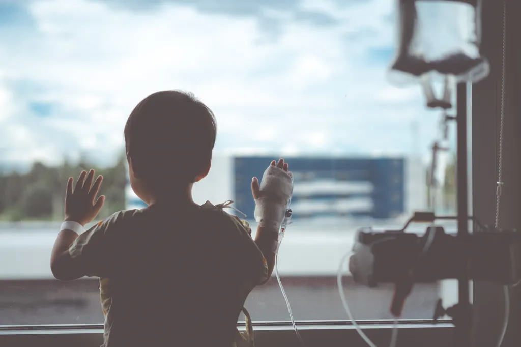 Toddler boy with IV tube looking out hospital window.