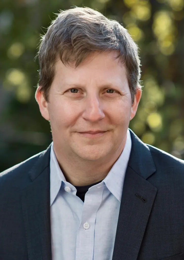 Headshot of a man with short brown hair, wearing a dark suit jacket and a light blue button-down shirt.