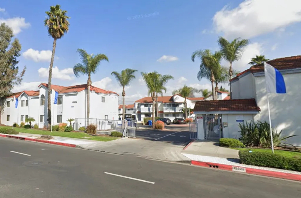 View of an apartment complex from the street, with white buildings, red tile roofs, palm trees, and an open gated entrance.