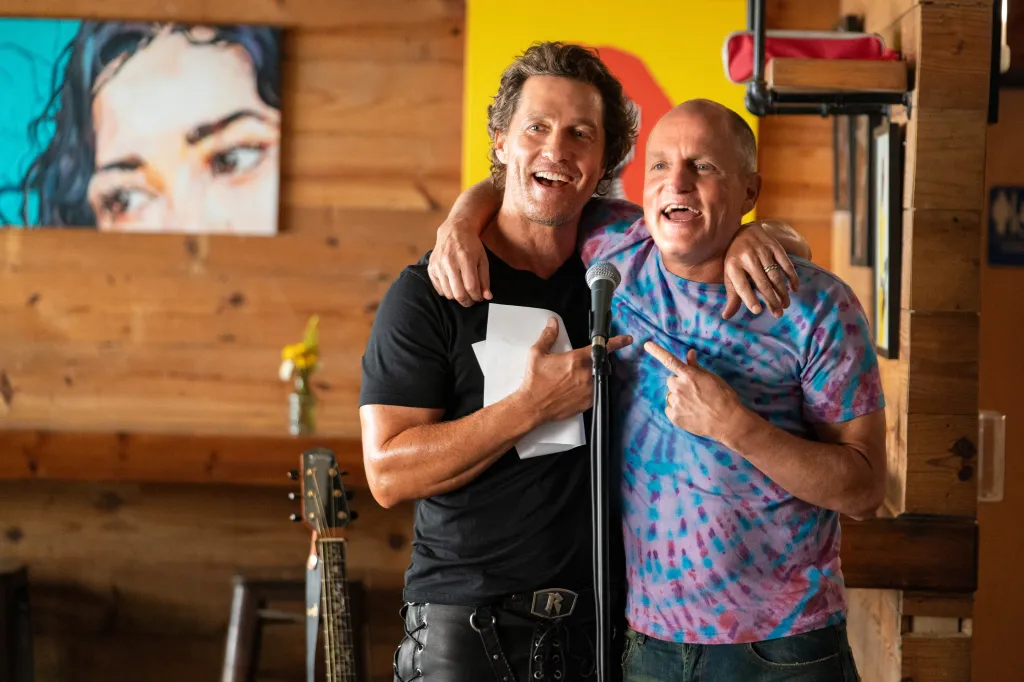 Matthew McConaughey and Woody Harrelson standing at a microphone, smiling with their arms around each other.
