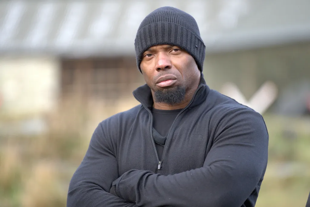 A man wearing a black beanie and a black zipped top looks directly at the viewer with his arms crossed.