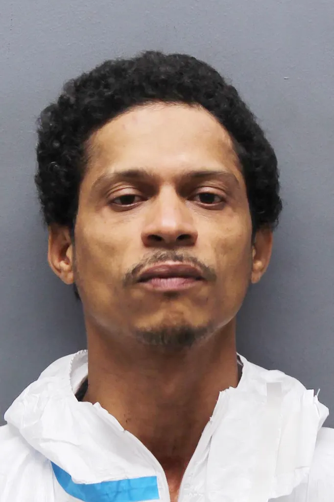 Mugshot of a man with dark curly hair and a mustache and goatee, wearing a white jumpsuit with blue trim.