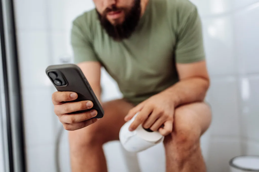 A person sitting on a toilet with a phone in one hand and a roll of toilet paper in the other.
