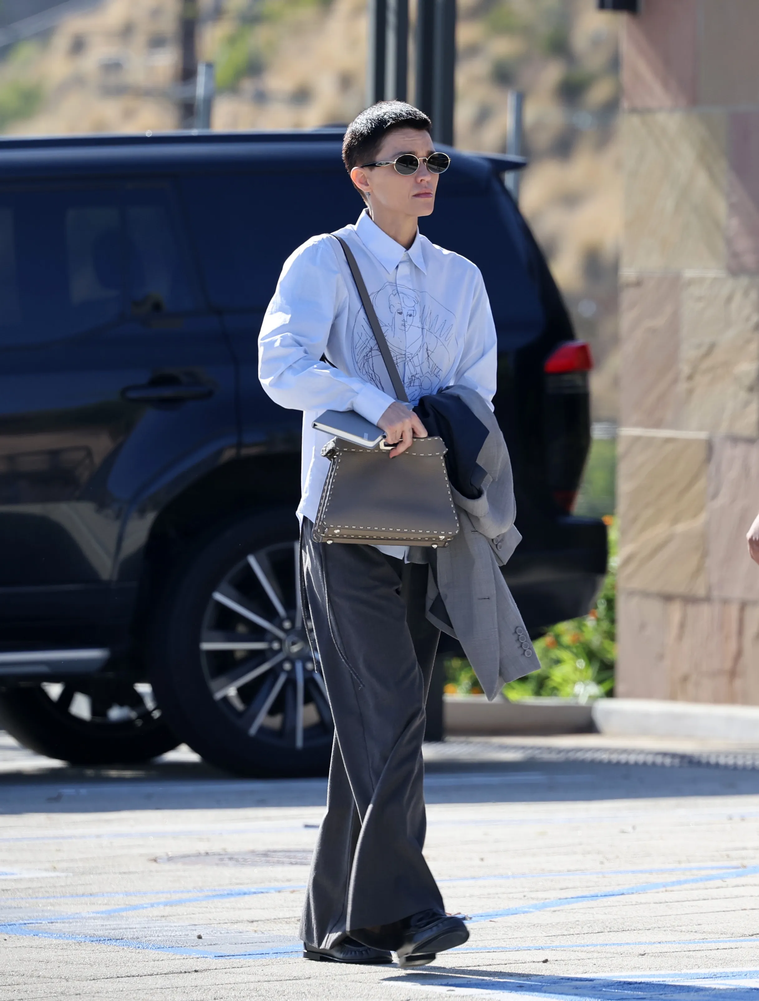 Ruby Rose walking outdoors, wearing a white shirt, gray wide-leg pants, and sunglasses.